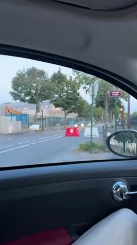 The video is taken from inside a car, looking out the passenger side window. The camera is pointed at a street with trees lining the sidewalk. In the foreground, part of the car's interior is visible, including the door panel, handle, and a portion of the passenger seat. The street is a two-lane road with white lane markings. On the road, there are several red and white temporary traffic barriers, some of which have signs. One sign on a red barrier is a blue circular sign with a white arrow pointing to the right, indicating a mandatory direction. A black car is driving down the street, approaching the barriers. A white traffic barrier is visible further down the road. On the sidewalk, there are trees, a lamppost, and a trash can. In the background, there are buildings and a fence. The sky is overcast.