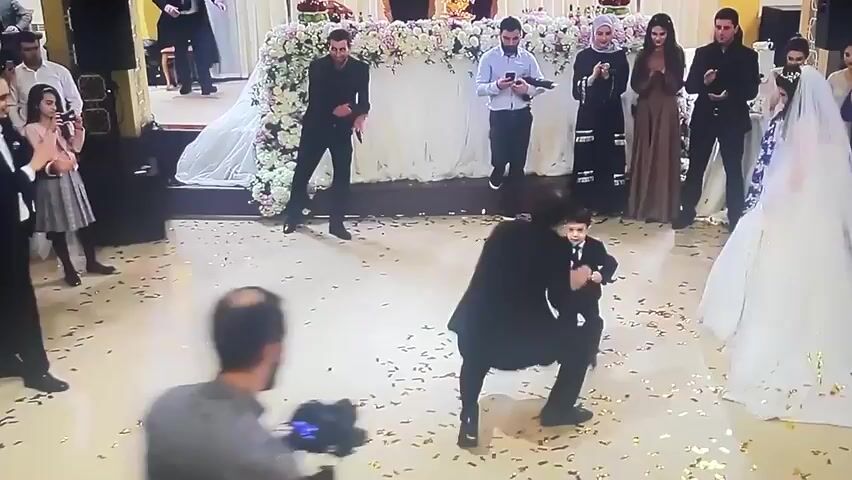 This video captures a wedding celebration. A young boy in a suit is enthusiastically dancing in the foreground, his movements energetic and playful. He is surrounded by a crowd of wedding guests who are watching him and clapping. To the right of the boy, a bride in a white wedding dress with a long veil is visible, appearing to be dancing or walking. Other wedding guests are dressed in formal attire, and some are holding cameras or phones, suggesting they are documenting the event. The environment appears to be a decorated indoor venue, possibly a banquet hall, with floral arrangements and ornate lighting. Confetti is scattered on the floor, indicating a festive atmosphere. The overall impression is one of joy and celebration at a wedding.