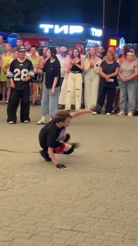 The video shows a young male breakdancer performing on a cobblestone street. He is wearing a black t-shirt with a white stripe, black shorts with red accents, and black kneepads. He is in the middle of a dynamic move, with one arm on the ground and his body in a twisted position. A crowd of people, mostly adults and teenagers, are standing behind him, watching the performance. They are dressed in casual summer clothing. In the background, there is a building with a neon sign that reads "ТИР" in large letters. The lighting suggests it is evening or night. The overall environment appears to be an outdoor public space, possibly a pedestrian walkway or promenade. The focus is on the breakdancer's athletic movement.