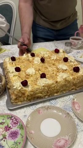 A man is cutting a square cake. The cake has white frosting, is sprinkled with crumbs, and is decorated with dollops of frosting and cherries. The cake is on a silver serving tray on a table covered with a white tablecloth. Several floral patterned plates are around the cake. A cup of tea, a small glass, and a salt shaker are also on the table. The man is wearing a t-shirt and jeans. Another person's arm is visible, and part of a chair is also visible.