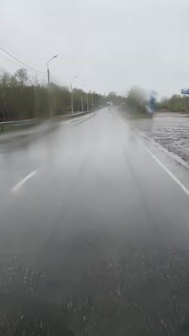 The video shows a rain-soaked asphalt road stretching forward under an overcast sky. The road is lined with bare trees and streetlights on either side. A few vehicles are visible in the distance, driving along the road. There is standing water on the sides of the road and the surface of the road is reflective from the rain. A metal guardrail is visible to the left of the road. The camera appears to be stationary.