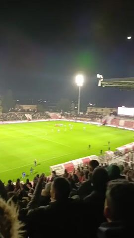 The video depicts a soccer game at night, viewed from the stands behind a team bench. The field is brightly lit by floodlights. Players in red and white uniforms and blue uniforms are positioned around the center of the field. Fans fill the stands, many wearing jackets or hoods. The sky is dark, with a visible moon. The perspective is from a spectator, providing a full view of the soccer field and the surrounding stadium, but primarily showing the spectators nearby.