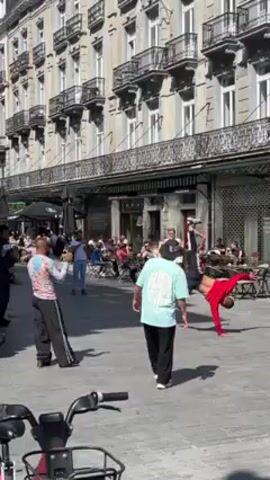 The video shows a street performance in front of a café. A man in a red outfit is performing a breakdancing routine. Several people are standing around watching the performance, while others are seated at tables outside the café. The background shows a tall building with many windows and balconies. There is a bicycle parked in the foreground, partially blocking the view. The scene is well-lit, suggesting it is daytime.