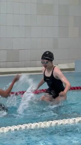 The video shows four young people in a swimming pool. Two individuals are in the foreground, one on the left and one on the right. The person on the right, wearing a black swimsuit and goggles, is holding a pink kickboard and appears to be laughing. They are sitting on the shoulders of another person, also wearing goggles. The person on the left is wearing a black cap and goggles and is reaching out towards the person on the right. There is a lot of splashing water around them. In the background, two more individuals are visible in the water. One is in the middle left, raising their arms. The other is in the middle right, partially submerged. The pool has lane lines and a tiled wall in the background. The overall scene depicts a playful and energetic atmosphere.