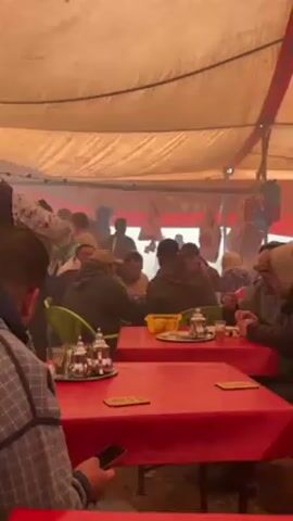 Here's a detailed description of the video:

The video shows the interior of a large tent-like structure filled with people sitting at tables, likely a restaurant or eating area. The tent's ceiling is a light-colored fabric, possibly canvas, with some red trim along the edges. The space is visibly smoky, creating a hazy atmosphere.

People are seated at red tables, most in green plastic chairs. Each table has a tea set, including a silver teapot and glasses. Individuals are either eating, drinking, or socializing. Several are wearing warm clothing like jackets and hats, suggesting a cold environment outside. One man, facing the camera, has a mustache and wears a dark beanie. Another man is looking at his phone.

In the background, more people are moving about, and there appear to be items hanging from the tent's structure. The overall impression is of a bustling, temporary gathering place, possibly at an outdoor market or event.