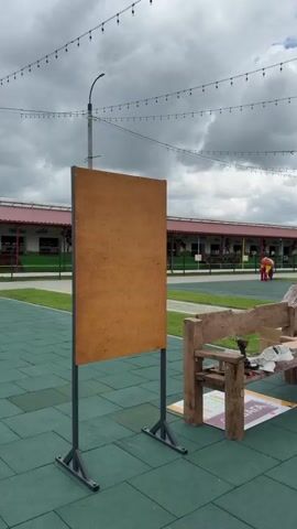 A short video pans across a children's outdoor play area. In the foreground, a rustic wooden bench is visible with tools and papers on it. Beyond the bench, a paved walkway leads through a grassy area. Further in the background, a row of stalls or buildings with red roofs can be seen. There are playground structures, including a trampoline and a large inflatable bounce house. String lights are strung across the sky. The sky is cloudy with patches of blue. People are visible in the distance, including a person sitting on a bench, a child running, and a person in a costume. The ground is covered in green rubber mats.