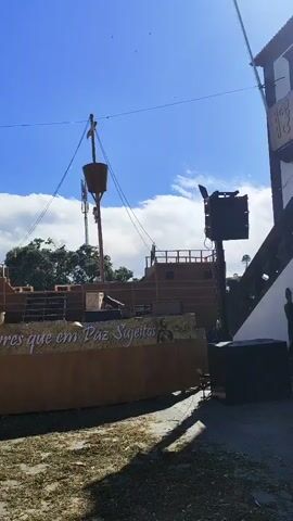 The video captures an outdoor scene with a replica of an old wooden ship as the central focus. The ship is decorated with what appears to be a banner or inscription that reads "Antes Morrer Livres que em Paz Sujeitas". The ship is situated on grassy ground, and in front of it, there is a large speaker system mounted on a pole, with additional speaker boxes at the base. To the left of the speaker system, a white Isuzu flatbed truck is parked. In the background, there are buildings, including one with white walls and a yellow-domed roof, and other structures that suggest a town or event space. The sky is blue with some scattered clouds, indicating a daytime setting, likely a fair or festival. There are some indistinct figures of people visible in the far background.