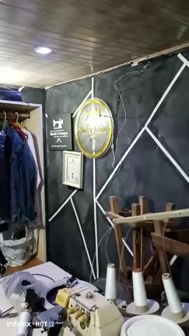 The camera pans across a dimly lit room. The ceiling is covered with wooden planks. The walls are painted dark gray and decorated with a geometric pattern of white stripes. The room appears to be a tailor's shop. There is a sewing machine on a table, along with spools of thread. Several overturned wooden chairs are also in the room. On the wall are two framed images, one with an emblem of a tailor and the other a certificate. There are two mannequins dressed in black strapless swimsuits. The video ends.