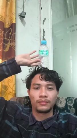 A person is sitting in a chair, with a plastic water bottle balanced on their head. A hand is shown reaching up towards the bottle, appearing to steady it. The person is wearing a plaid shirt. The background shows a curtain and a wall.