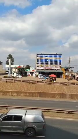 The video shows a view from across a highway, with a building and a gas station on the other side. The building is called "SELFRIDGES PIONEER" and has a blue window facade. Next to it, there is a gas station, and people are walking in front of the building and around some red umbrellas. A yellow truck and a gray truck pass by on the highway in the foreground. The sky is blue with white clouds.
