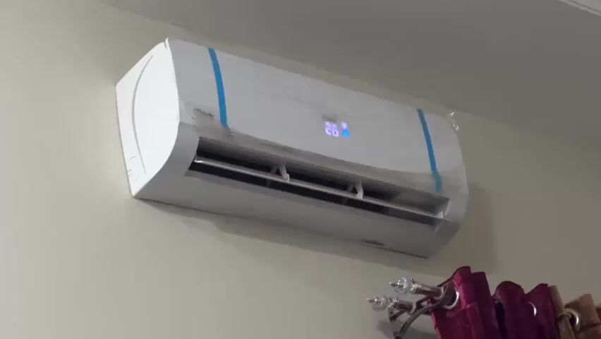 The video shows a split air conditioner mounted on a light-colored wall. The air conditioner is white with blue decorative tape on its sides. A digital display on the front of the unit shows the temperature as 26 degrees. Below the air conditioner, a portion of a curtain rod with purple curtains is visible. The lighting in the room appears to be artificial.