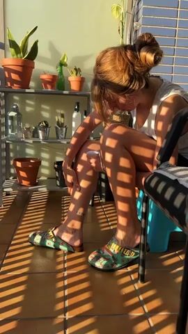 A young woman with auburn hair in a bun sits on a chair on a balcony. She is wearing a grey t-shirt and colorful slip-on shoes. Sunlight streams through a window, casting striped shadows across her legs and the tiled floor. On a shelf behind her are several potted plants, a bottle, and other decorative items. The woman is looking down at her leg and appears to be touching it.