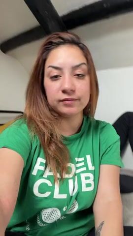 The video shows a woman wearing a green t-shirt with the words "Padel Club" printed on it. She is looking at the camera and speaking. In the background, there are two people lying down. One person is wearing a yellow hoodie and the other is wearing a black t-shirt. They appear to be on a couch or a similar piece of furniture. The lighting suggests an indoor setting.