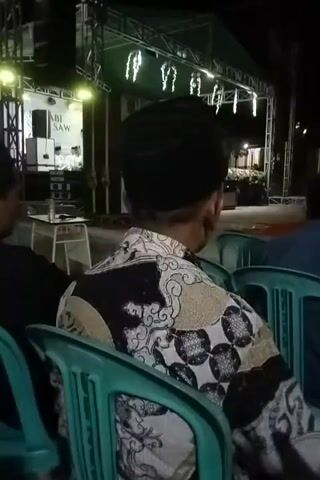 A video clip begins with a close-up shot of the back of a person wearing a black peci hat and a patterned shirt, seated in a green plastic chair. The camera slowly zooms out to reveal a stage in the background, decorated with lights and a banner that reads "NABI SAW". A sound system is visible on the stage. The scene appears to be an outdoor event at night. Another person is partially visible to the left of the main subject, also seated. The overall ambiance suggests a performance or gathering.
