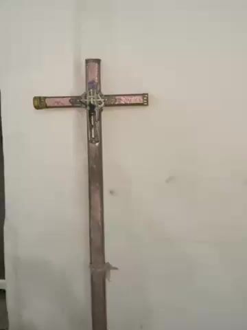 The video shows a standing cross with a decorative emblem at the intersection of the vertical and horizontal beams. The cross is affixed to a white wall. The cross itself appears to be metallic or painted with a metallic finish in a color reminiscent of rose gold or copper. The emblem in the center of the cross features ornate detailing and may have a smaller cross or figure within it. The room appears to be sparsely decorated with a plain wall and minimal other visual elements. There are no people visible in the video.