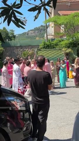 The scene captures a joyous celebration on a sunny day, likely a wedding or pre-wedding event, taking place on an outdoor street with buildings and a distant mountain in the background. The focus is on a crowd of people, many of whom are dressed in vibrant, traditional Indian attire, including pink kurtas and saris. At the center, individuals are dancing and hugging, their movements animated and lively. Several people, presumably guests or observers, are watching the festivities, and one person is filming the event with their phone. A dark car is parked in the foreground, partially obscuring the view, while a leafy tree branch overhead frames the scene. The overall atmosphere is festive and cheerful.