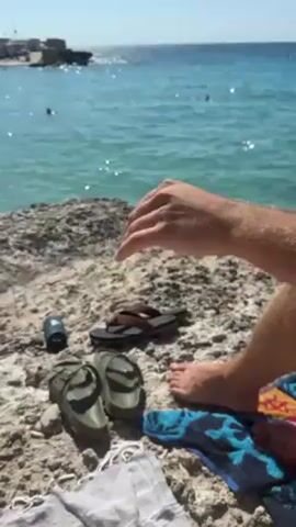 The video shows a man relaxing on a rocky beach. His bare feet are visible, and a few pairs of sandals and a towel are placed next to him on the rocky ground. A small speaker sits near the sandals, playing music. In the background, the clear turquoise water of the sea sparkles in the sunlight, and a few people can be seen swimming in the distance. The sky is clear and blue.
