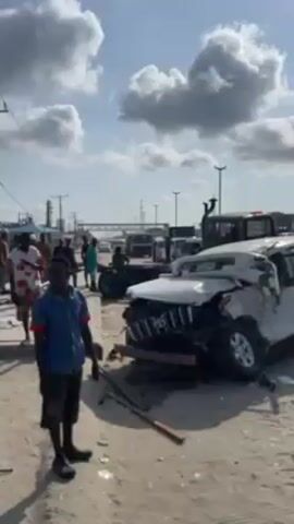 The video shows the aftermath of a car accident. A white Toyota Prado is severely damaged in the front. Several people are standing around the vehicle and a tow truck is visible in the background. The scene is taking place outdoors on a sandy ground area. There are other vehicles parked nearby, including what appears to be a police vehicle. The sky is partly cloudy.