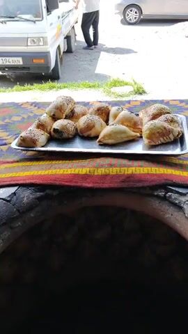 The video shows a tray of freshly baked samsa pastries, some triangular and some round, sprinkled with sesame and black seeds, placed on a colorful embroidered cloth. The cloth is laid out in front of a large, circular clay oven (tandoor), indicating a traditional baking method. In the background, a person is visible near the back of a white pickup truck, and a silver car is parked further away. The overall scene suggests an outdoor food preparation or market setting, with an emphasis on the appetizing samsa pastries.