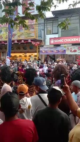 The video shows a street performance with a large crowd gathered. In the foreground, the backs of several people are visible, some holding up phones to record the event. The crowd is dense and appears to be watching something happening in the center of the street. There are performers in the middle wearing elaborate costumes, some with large, furry wigs and masked faces. Behind the performers, there are shops and buildings lining the street, including a "SONIC CHICKEN" restaurant. The atmosphere seems festive and crowded, with people in various attire, including some wearing headscarves. The time stamp indicates the video is approximately four seconds long.
