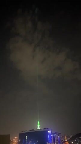 A green laser beam originates from the apex of a illuminated building and extends upwards into the dark, cloudy sky. The beam appears to slightly diffuse as it ascends, and there are some faint green lights or reflections visible higher in the frame, possibly within or above the clouds. The building is modern, with blue lights illuminating its facade and possibly some signage. The overall scene is nocturnal, with a city skyline dimly visible in the background.