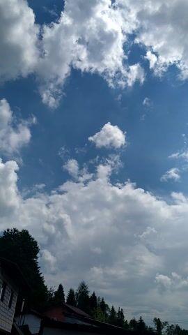 A time-lapse video shows the sky with clouds moving. The sky is a bright blue with white clouds scattered throughout. Some of the clouds are large and fluffy, while others are smaller and more wispy. In the lower right corner of the frame, there are some trees and buildings visible. The video is shot from a low angle, looking up at the sky. The clouds are moving quickly, and the sun is shining brightly through them. The overall effect is one of a dynamic and ever-changing sky.