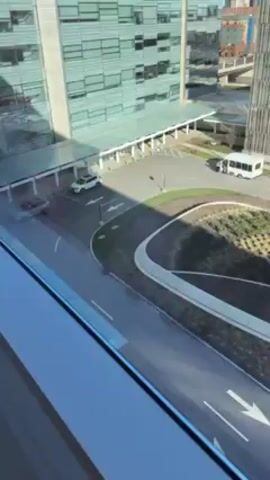 The video shows an elevated view of a street scene from a window. In the foreground there is a round-about type island with greenery and bare trees. The street has white directional arrows. A white bus moves on the street as a small red vehicle moves along the cross walk. Behind the street is a large parking garage with a yellow entrance and logo. People are walking on the sidewalks. The sky is bright and there are several tall buildings in the background.