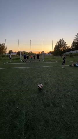 A group of young people are playing soccer on a field at sunset. The sun is setting behind a goal, casting a warm glow over the scene. There are several people in the background, some standing in front of the goal as if defending it, and others are on the field. One person is in the foreground, about to kick a soccer ball. The field appears to be artificial turf. The overall mood seems to be one of fun and recreation, with the players enjoying the late evening light.