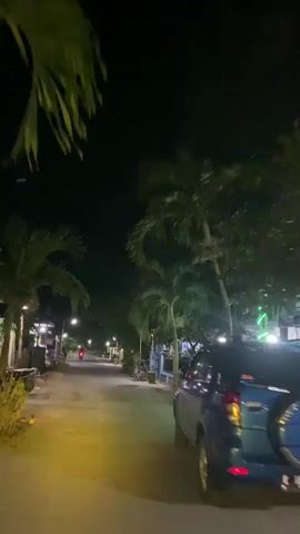 The video captures a nighttime view of a quiet street lined with palm trees and buildings. Streetlights illuminate the asphalt road, casting a warm glow. A few parked motorcycles are visible on the side of the road. In the distance, a red light from a moving vehicle can be seen. The sky is dark, indicating it is late at night or early morning. A blue vehicle is partially visible on the right side of the frame. The overall atmosphere is calm and peaceful.