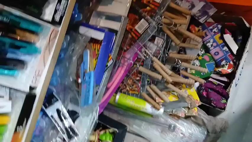 The video shows a messy glass display case in what appears to be a shop. The case contains a variety of items including toiletries, small toys, candy bars, makeup items, and other assorted merchandise. The items are scattered haphazardly, with a lot of plastic packaging visible. The camera moves slightly, showing more of the display case. Some shelves behind the glass are also visible, stocked with more items. There are no people visible in the video.