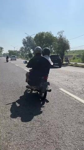 The media file shows two people riding a motorcycle on a sunny day. They are both wearing helmets and are facing away from the camera. The motorcycle is dark-colored and appears to be a scooter. The scene is a paved road with traffic, including cars and other motorcycles. Trees line the side of the road, and a hill with vegetation is visible in the background. The sky is clear and blue, indicating good weather. The time of day appears to be late morning or early afternoon, given the bright sunlight and the length of the shadows. The overall atmosphere seems peaceful and ordinary, capturing a common mode of transportation in the depicted location. No specific emotions are discernible from the subjects due to their obscured faces. The license plate on the motorcycle is partially visible, with the numbers "2302" discernable. The road appears to be in a rural or semi-urban setting, possibly in or around Kedungbendo, Indonesia, as per the provided location context.