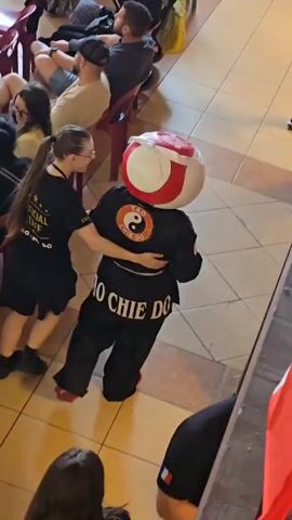 Here's a concise description of the video:

The video shows a person in a martial arts uniform and a large, spherical head costume.  The uniform is black with "VO CHIE DO" printed on the back in white letters. The head costume is white with red accents and a yin-yang symbol on the back.  The person is being guided by a woman in a black t-shirt and shorts with her arm around their waist. They are walking past a seated audience in an indoor space with tile flooring. Some people are watching the scene, and others are looking at their phones or talking. Flags of the country of Belgium are visible.