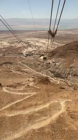 The video begins with a view looking down a steep, rocky mountainside with a winding path visible. A cable car is suspended from wires overhead, moving away from the viewer and towards a structure on the mountainside. In the distance, a body of water, possibly a lake or sea, is visible under a clear blue sky. The landscape is arid and appears to be a desert. The cable car continues its descent, offering a progressively wider view of the winding path and the vast, dry terrain. The image is stable, suggesting it was filmed from inside the moving cable car.