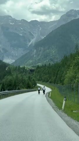 The video shows a gray, overcast day with a winding asphalt road leading through a mountainous, forested valley. The road appears to be paved and is in good condition, with a metal guardrail on the left side and a wire fence on the right. The road curves to the right as it goes deeper into the valley.

In the foreground, two people are cycling away from the camera on bicycles. They are wearing casual clothing. The cyclist on the left is wearing a white top and dark shorts, while the cyclist on the right is wearing a dark top and dark shorts. They are riding side-by-side.

The surrounding environment is dominated by lush green pine trees that cover the steep slopes of the mountains. The mountains themselves are rugged and imposing, with some bare rock faces visible at higher elevations. The sky is heavily clouded, suggesting the possibility of rain. In the distance, a dark structure, possibly a tunnel entrance, is visible nestled within the trees on the left side of the road. There are also some small, indistinct animals grazing on the grassy area to the right of the road. The overall impression is of a serene and somewhat wild natural landscape, with the cyclists adding a human element to the scene.