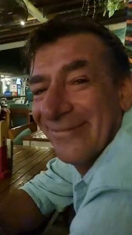 A man is seated at a wooden table at an outdoor restaurant. He is wearing a light blue button-down shirt. He squints his eyes and smiles, reacting to something just outside of the frame. A man in a turquoise shirt is visible in the background, along with several other blurred people. The restaurant has a wooden ceiling and appears to be in a tropical location, with palm fronds near the ceiling.