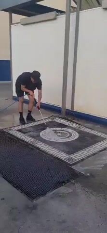 A person wearing a black t-shirt, black shorts, black socks, and black shoes, with a black cap, is pressure washing a rug. The rug is dark with a geometric pattern in shades of white and gray, featuring a circular floral design in the center. The person is holding a pressure washer wand and directing the spray onto the rug, which is lying on a concrete surface. Water and suds are visible on the rug and the surrounding ground. A metal grate is to the left of the rug. The background consists of a white wall with blue accents and vertical support beams. The action appears to be taking place outdoors in daylight.
