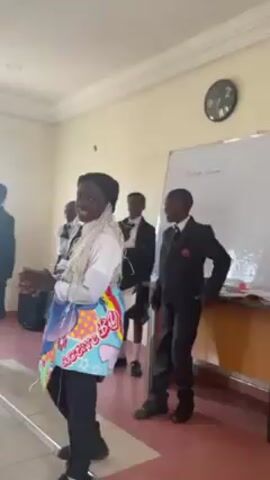 The video shows a group of school children, mostly dressed in formal uniforms, in what appears to be a classroom setting. The prominent figure in the foreground is a student wearing a beanie, a white shirt, black pants, and what appears to be a colorful, printed apron or sash. They are actively engaged, gesturing with their hands and speaking. Behind them, other students stand or lean against furniture, seemingly observing or waiting for their turn. A whiteboard is visible in the background with the words "Black Drama" written on it. There is a wall clock and some school furniture in the classroom as well.
