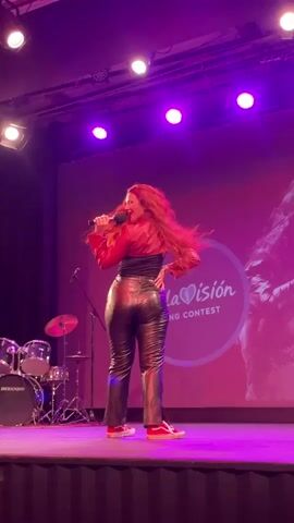 A woman with long, wavy red hair is performing on a stage. She is wearing a red jacket, a black top, and shiny black pants, along with red sneakers. She is holding a microphone and appears to be singing and dancing. The stage is lit with purple lights. Behind her is a large screen displaying a logo that includes the words "Aula" and "Song Contest", along with a heart symbol. To the left of the stage, a drum set is visible. The woman's back is to the camera initially, and she then turns to face forward as she sings and gestures.