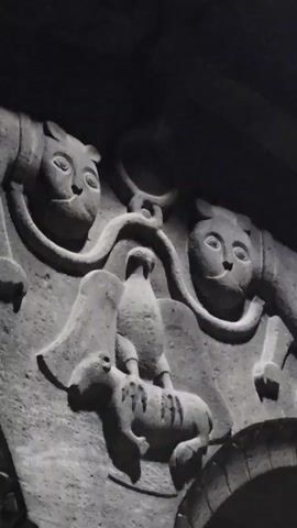 The video shows a close-up of a stone carving. The carving depicts a scene with several animals. On the left and right sides of the carving are two stylized animal heads that resemble cats or lions. Between these two heads, a chain or rope hangs down, attaching to a ring at the top. Below the ring, there is a central carving of a bird standing on the back of a quadruped animal, possibly a sheep or a dog. The bird is facing forward, and the quadruped is facing left. The entire carving is set into a stone facade, with arched openings visible at the bottom of the frame. The lighting is dim, with shadows highlighting the details of the carving.