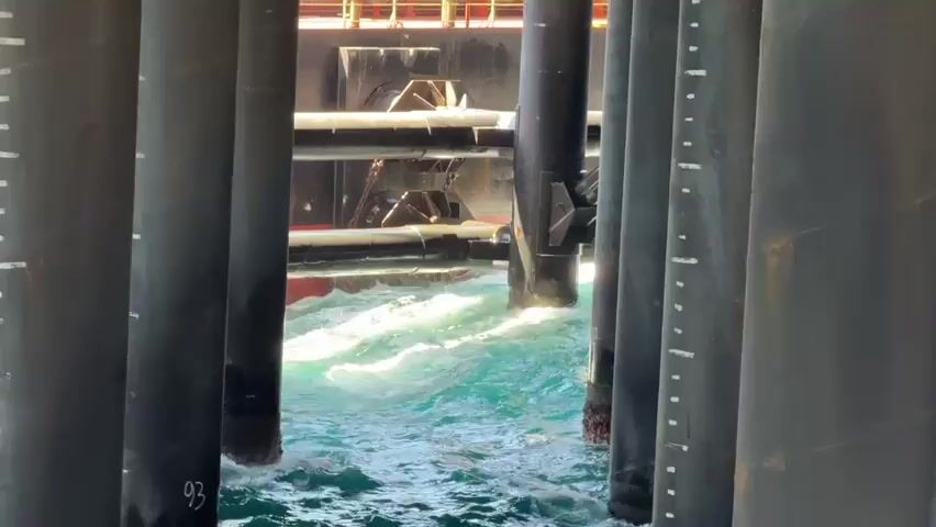 The video shows a choppy, turquoise sea viewed from underneath a series of large, black, cylindrical structures. The structures appear to be part of a pier or a similar offshore installation, with white markings indicating measurements along their sides. Between these structures, a large, dark vessel is visible. Its hull is red and black, and it has orange railings on its upper decks. A horizontal pipe or beam extends from the vessel, and it is submerged in the water, creating splashes and turbulent waves around it. The water is rough, with white foam and spray. The perspective is from below the water level, looking out towards the vessel.