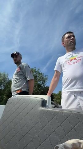 Two men are on a boat on a sunny day. The man on the left is wearing a gray t-shirt and a baseball cap. He is looking to the left. The man on the right is wearing a white t-shirt and white shorts. He is looking forward. The boat has gray seats. There are trees in the background and a blue sky with clouds. The man on the left puts his hand on the boat's railing.