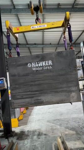 A large, gray, rectangular object bearing the text "HAWKER Water Less" is suspended from a yellow lifting beam by four purple straps. The beam, which has a working load limit (WLL) of 3,200 KG, is attached to a hook hanging from above. The scene appears to be inside a large industrial building with a high, metal ceiling. To the right of the suspended object, a portion of a forklift is visible. The flooring is concrete and various pieces of metal equipment and supports are visible in the background.