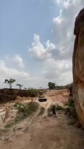 The video shows a bulldozer moving on a dirt road, on a construction site or quarry under a bright blue sky with fluffy white clouds. There are at least two children visible, one sitting against a rock wall and one running along the road. The road and surrounding land are mostly dirt and gravel. There are also some green trees and bushes in the background.