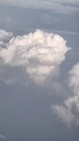 The video shows an aerial view of clouds. A large, puffy white cloud dominates the center of the frame, with a smaller, elongated white cloud to its right. The background consists of a hazy, muted blue sky with other indistinct clouds. The camera appears to be moving, as evidenced by the slight shift in perspective and the apparent motion of the clouds.