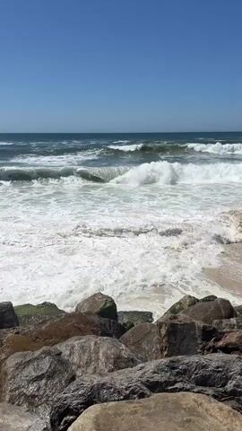 The video shows a rocky shoreline with waves crashing on a beach. The ocean is a deep blue, and the sky is clear and bright, indicating a sunny day. The waves are powerful, with white foam cresting as they break. In the foreground, large rocks of various sizes and colors are arranged along the shore, some covered in green moss. The overall scene evokes a sense of natural beauty and the raw power of the ocean. The time of day appears to be midday, given the bright sunlight. The location is likely Torreira, Portugal, as indicated by the context. There are no people or man-made objects visible in the frame, focusing solely on the natural elements of the seascape. The video captures the dynamic movement of the water and the rugged texture of the rocks.