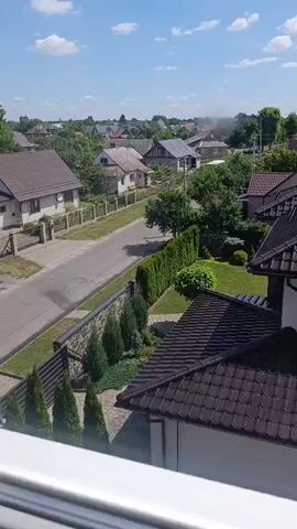 The video shows an aerial view of a residential neighborhood on a clear day. The scene is dominated by houses with brown tiled roofs and well-maintained gardens. A paved road runs through the middle of the frame, lined with fences and hedges. 

In the foreground, the roof of a large house with a dark brown tiled roof is prominent. Beyond this, a row of cypress trees lines a stone wall, separating the property from the road. Further down the road, several other houses are visible, each with its own garden and fence. The background reveals a more distant cluster of houses and trees, suggesting a suburban or semi-rural setting.

The time of day appears to be daytime, with bright sunlight casting shadows, indicating a sunny weather. The sky is blue with a few scattered white clouds. The overall atmosphere is peaceful and serene. There is no visible text in the media file.