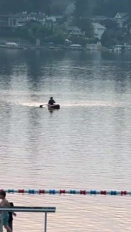 A person on a paddleboard is in the middle of a lake. The person is facing left and is holding a paddle. The water is calm and there is a slight haze in the air. In the background, there are houses and trees on the shore. In the foreground, there is a rope with red and white buoys.