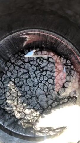The video shows a close-up, top-down view of a pot filled with a dark, viscous liquid. The surface of the liquid is covered in a cracked, textured pattern, resembling dried mud or a cracked earth effect. There are reflections on the surface, indicating a light source, possibly sunlight, casting a warm glow on the right side of the frame. The liquid appears to be thick and possibly boiling or cooling, as indicated by the bubbling or cracking texture. The pot itself is a dark, metallic color, suggesting it's a cooking pot. There are no people visible, and the setting appears to be outdoors due to the strong natural lighting. The overall mood is somewhat abstract and focuses on the mesmerizing texture and movement of the liquid. The time of day appears to be daytime, likely sunny, given the intensity of the light and reflections. There is no visible text in the media.