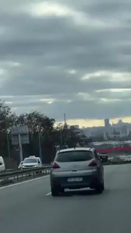 A car travels on a highway under a cloudy sky, with the Eiffel Tower visible in the distance.  The highway has multiple lanes and the car is in the right lane.  Other vehicles, including a truck, a white car, and a motorcycle, are also on the highway. The surrounding landscape includes trees and city buildings.  The sky is overcast with grey clouds.