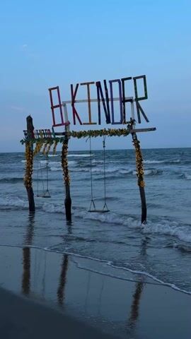 The video presents a stationary shot of a beach scene featuring a wooden structure with swings and the word "AKINDER" spelled out in colorful letters atop it. The structure is adorned with artificial sunflowers and sits partially submerged in the ocean waves. The water is choppy, and the sky is a clear blue. The overall ambiance is calm and serene, highlighting the juxtaposition of the man-made structure with the natural environment of the sea.